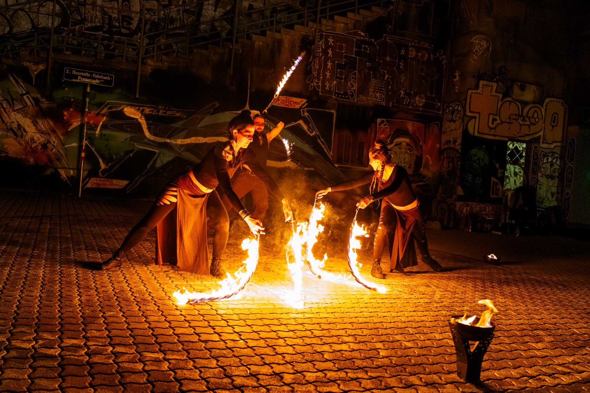 Drei Feuerkünstler:innen mit Feuerseilen und Stab vor einer Graffiti-Wand