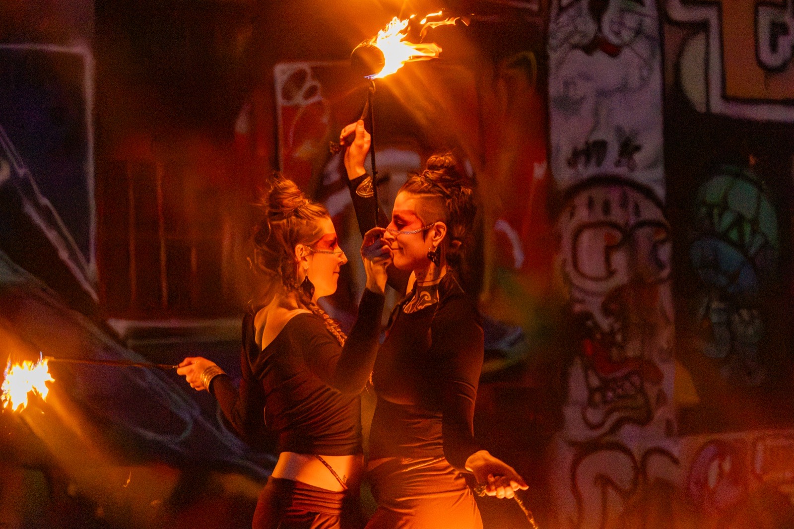 Zwei Feuerkünstlerinnen mit brennender Requisite vor Graffitiwand bei Nacht